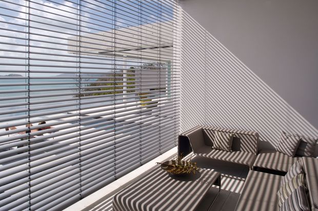 Bright contemporary living area beside extensive horizontal venetian blinds, with strong striped sunlight and shadow patterns across a neutral sofa, ottoman-style coffee table, and walls. The glazing overlooks an urban/coastal setting, while the room has a clean, minimalist fit-out.