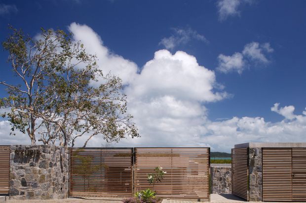 Exterior view of a contemporary property boundary with dark natural-stone masonry walls and horizontal timber privacy screens or gates. A sparsely branched tree, paved foreground, and distant landscape sit beneath a vivid blue sky with large white clouds.