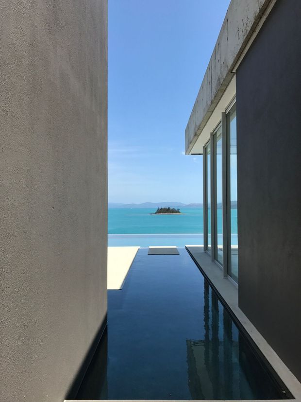 Narrow architectural exterior passage between smooth rendered walls, opening onto a turquoise waterfront view with a small island and distant hills. A long, dark reflective linear pool or water feature runs alongside full-height glazing beneath a projecting flat concrete roof. The composition is minimalist and strongly oriented toward the coastal outlook.