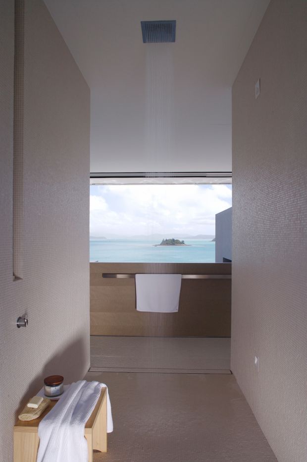 A minimalist bathroom with a freestanding bathtub partially visible in the foreground, a towel rail and folded towels, textured pale wall finishes, and a wide opening framing a turquoise ocean and small island beyond. The space is narrow and calm, with clean lines and a large scenic view.