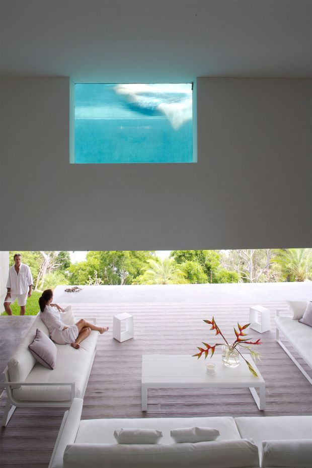 A bright, minimalist open-plan living area with white sofas, a low rectangular coffee table, and pale horizontal timber decking or flooring. The room opens to lush tropical landscaping, while a large rectangular aquarium-like underwater window or pool view is set high in the plain white wall above. The palette is predominantly white and light grey with turquoise blue and green outdoor accents.