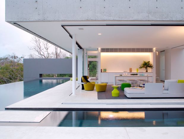 A sleek open-plan modern living and dining area opening directly onto a long reflecting pool and infinity-style swimming pool. The space has extensive floor-to-ceiling glazing, white walls and built-in cabinetry, a broad concrete overhanging roof, and minimalist white seating with vivid yellow and green accent chairs and stools. Warm concealed lighting illuminates the kitchen and dining zone, while landscaped trees are visible beyond the pool.