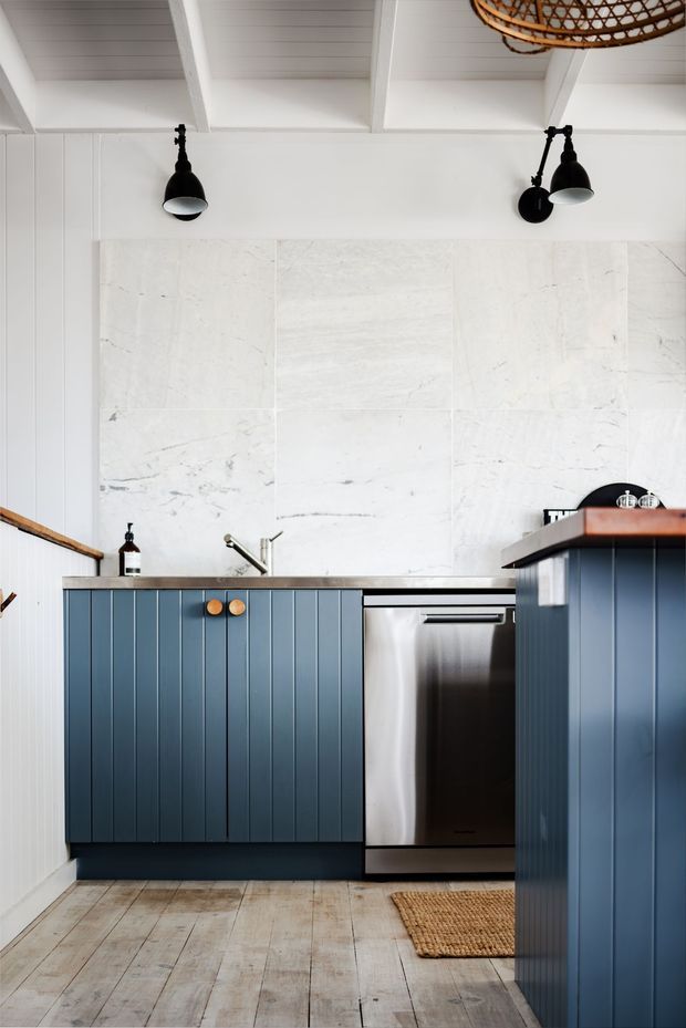 A compact, refined kitchen with deep blue vertical-panel cabinetry, a stainless-steel dishwasher, dark benchtops, and a large pale veined stone backsplash. White painted wall panelling and exposed ceiling beams create a bright coastal character, complemented by black wall-mounted lights, timber flooring, and woven accents.