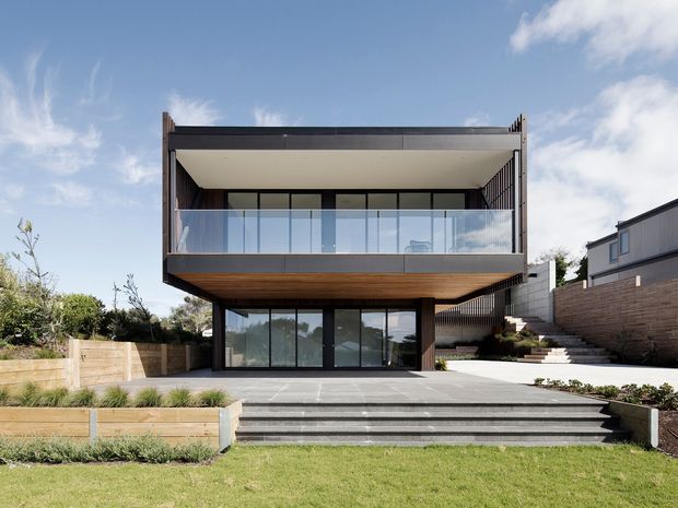 Contemporary two-storey house viewed from the rear garden, featuring a strongly cantilevered upper level with a deep covered balcony, frameless glass balustrade, dark horizontal cladding, extensive floor-to-ceiling glazing, and a broad stepped terrace leading to the lawn. The property sits on a landscaped, slightly sloped suburban site with timber retaining walls and neighbouring housing visible at the side.