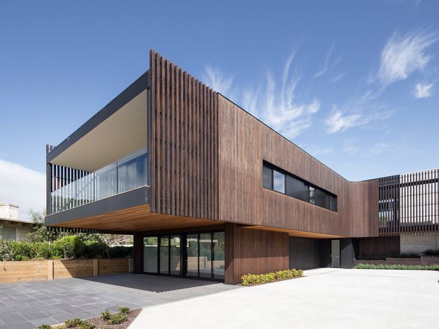 Exterior view of a contemporary multi-level residence with a strong rectangular form, dark timber vertical-screen cladding, projecting upper-floor volumes, extensive glazing, and a sheltered ground-floor entry or garage-like opening. The building sits beside a paved driveway and landscaped garden areas under a clear sky.