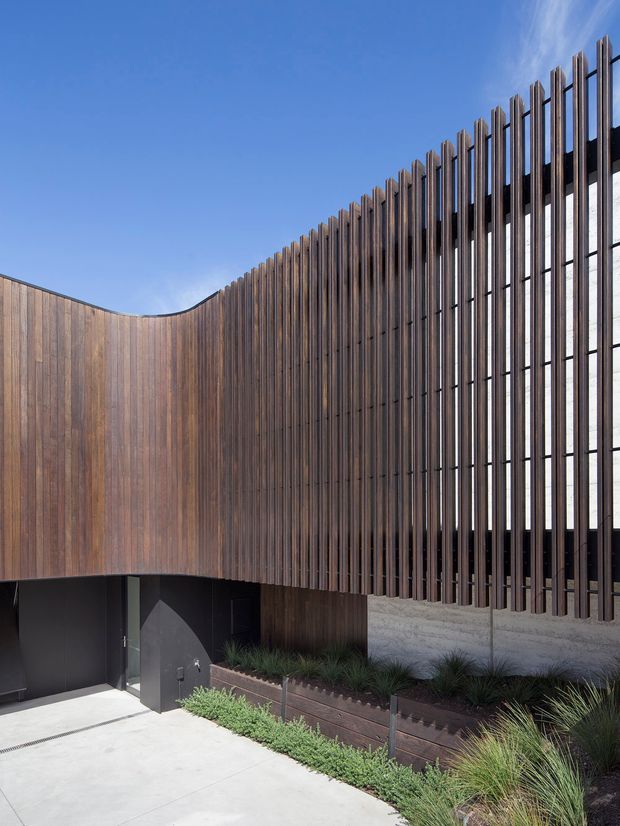 Exterior architectural view of a contemporary building with a sculptural curved facade. Vertical timber cladding wraps the left volume, while closely spaced dark vertical fins screen tall glazing on the right. A dark recessed entry, concrete paving, and a narrow landscaped planter with grasses are visible under a clear blue sky.