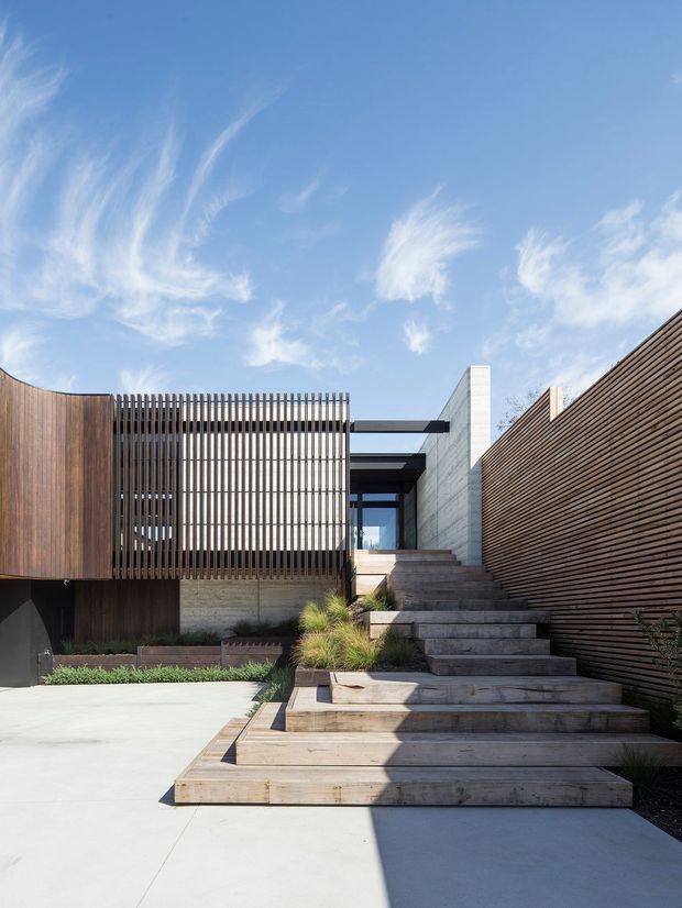 Exterior view of a contemporary residential building entry, featuring broad timber-clad stairs leading from a pale concrete forecourt to a recessed entrance. Vertical timber screening, horizontal timber slat fencing, exposed concrete walls, and dark steel canopy beams create a layered, geometric facade. The scene is brightly sunlit with a clear blue sky and sparse architectural landscaping.