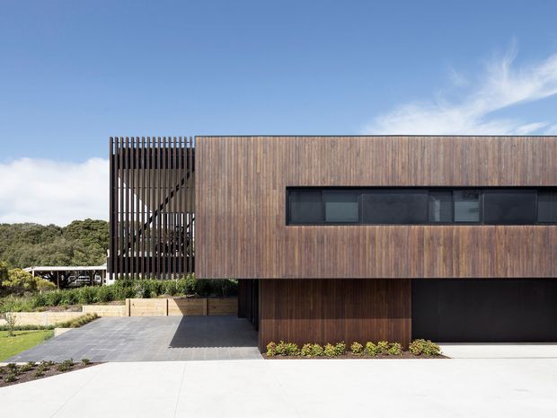Modern two-storey residential facade clad in vertical timber, with a long horizontal strip of dark glazing, a screened upper-level balcony or stairwell, and a dark garage opening. The house sits on a landscaped suburban site with a broad paved driveway and strong horizontal and vertical geometric lines.