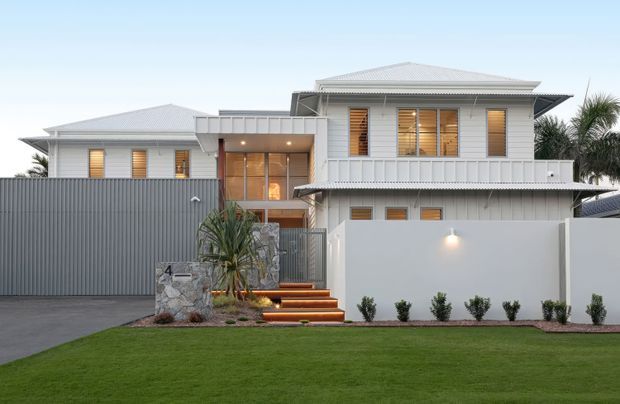Front exterior view of a contemporary multi-level residence with pale grey horizontal cladding, multiple low-pitched hipped roofs, timber-framed windows with warm blinds, a covered entry, rendered boundary walls, a ribbed metal side fence, stone feature wall and landscaped lawn.
