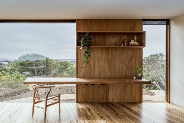A bright, minimalist interior featuring a built-in timber study desk and full-height shelving cabinet set against expansive glazing. A simple wooden chair sits at the desk, while the windows overlook landscaped terrain and a distant coastal-looking horizon. The room has polished timber flooring, white walls and ceiling, and warm natural wood joinery.