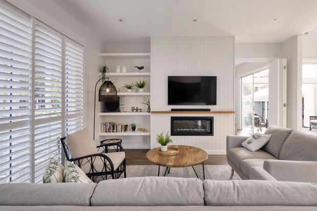 Bright, contemporary living room with a large light-grey sectional sofa arranged around a round timber coffee table. A built-in fireplace and television form the focal wall, finished with vertical light-toned panelling and a timber mantel. White open shelving displays books and plants, while full-height plantation shutters provide abundant filtered daylight. A glazed door opens to an outdoor area, visible in the background.