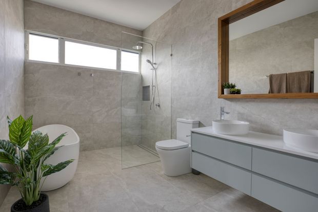 Contemporary bathroom with full-height pale stone-look tile across the walls and floor, a frameless glass walk-in shower, freestanding white bathtub, toilet, and a long floating grey vanity with two white vessel basins. A large timber-framed mirrored cabinet adds warmth, while clerestory-style windows provide natural light and indoor plants add greenery.