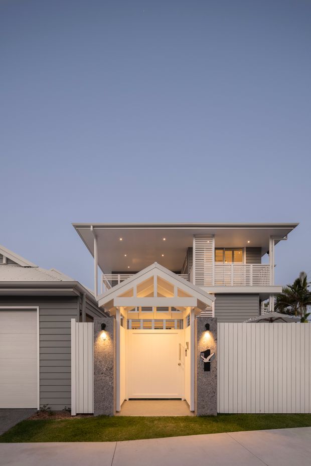 Twilight front exterior of a contemporary multi-level residence with a strongly defined covered entry. The façade combines grey horizontal cladding, white privacy fencing, a broad low-pitched roof with deep eaves, upper-level louvres and balcony screening, and a lit white gabled entry canopy over a tall gate. Warm recessed and wall lighting highlights the entry, with neighbouring rooflines and a palm visible at the edges.