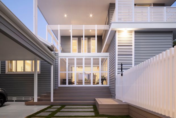 Exterior view of a modern multi-level residential development with dark horizontal cladding, extensive white louvres and privacy screens, balconies, external stairs, and a covered carport. Paved stepping stones and narrow lawn strips lead toward the entry, while warm interior lighting is visible through the screened glazing.