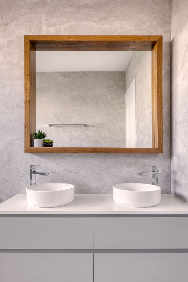 A contemporary double-vanity bathroom with two white vessel basins on a sleek wall-mounted vanity. The walls are clad in large-format grey textured tiles, complemented by chrome wall-mounted taps and a large rustic timber-framed mirror. Minimal decorative plants sit on the vanity beside the mirror.