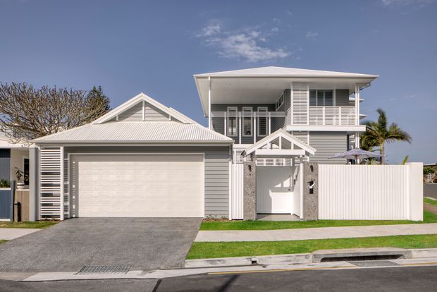 Street-facing exterior view of a contemporary two-storey coastal-style residence with light grey horizontal cladding, white trim, a low-pitched roof, upper-level balcony with white horizontal balustrades, and an attached single-car garage. The property has a paved driveway, white fencing, and a neat front lawn.