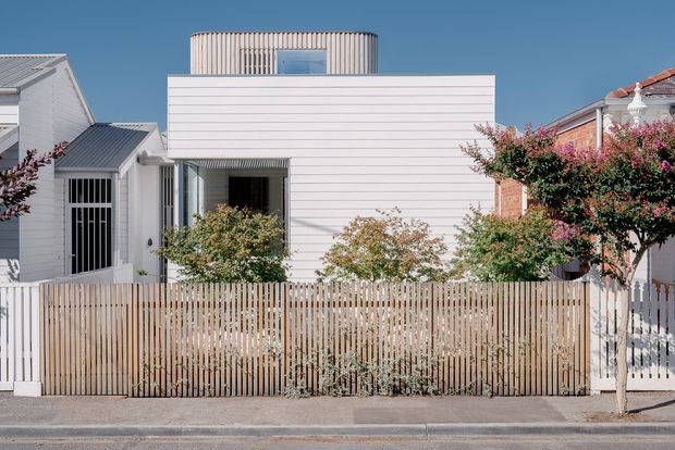 Street-facing view of a compact contemporary residence in an established urban neighbourhood. The house has a crisp white horizontal weatherboard facade, a projecting upper-level box with a cylindrical or rounded rooftop volume clad in narrow vertical boards, and a timber slat front fence. Mature ornamental trees and neighbouring homes frame the facade.