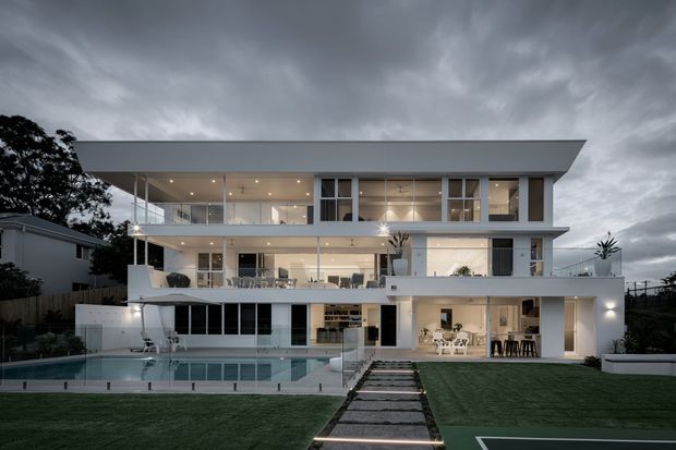 A large contemporary multi-storey residence viewed from the rear, featuring broad horizontal roof overhangs, extensive glazing, white rendered surfaces, open terraces, a swimming pool, landscaped lawn, and a lit stepping-stone path. The dusk setting and interior lighting create a polished, dramatic atmosphere.