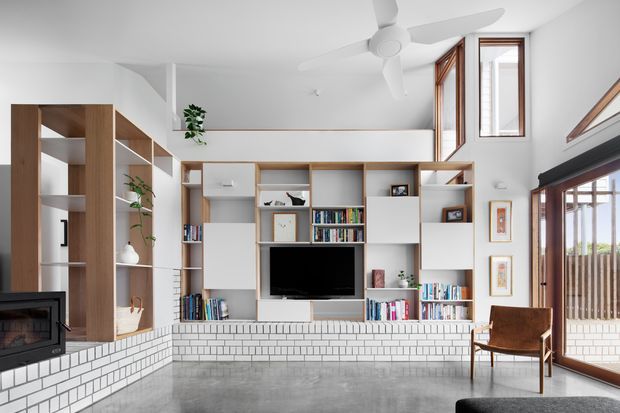 Bright contemporary living room with a large custom timber-and-white built-in shelving and media wall, exposed white brick fireplace surround, polished grey concrete floor, and extensive timber-framed glazing and doors opening toward an outdoor area. A ceiling fan, armchair, plants, books, and decorative objects furnish the open, airy space.