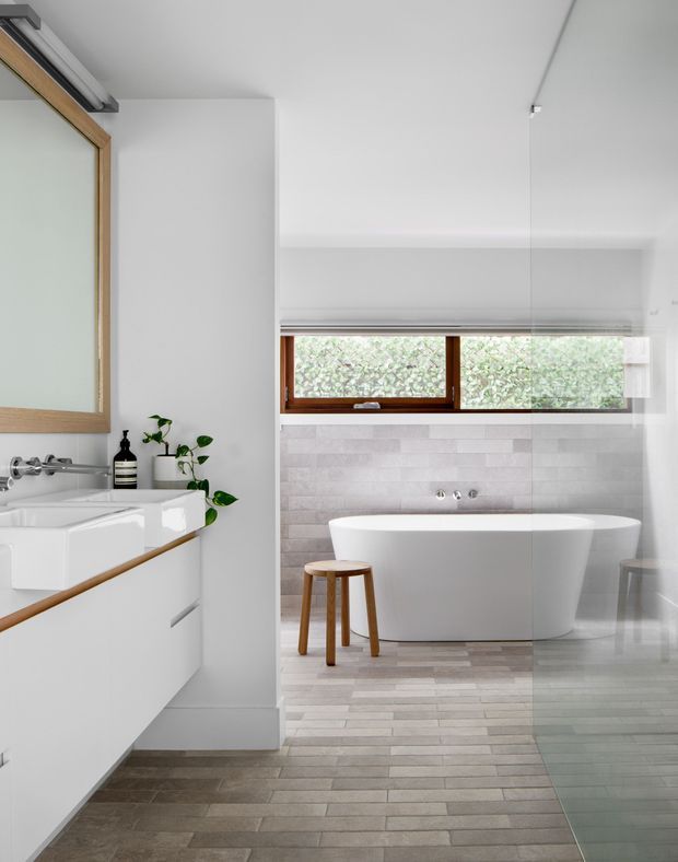 Bright contemporary bathroom featuring a freestanding white bathtub beneath a wide timber-framed window, a floating white vanity with integrated basin, pale tiled walls and floor, and warm timber accents. A glass shower enclosure is visible at right, with plants and simple toiletries adding natural detail.