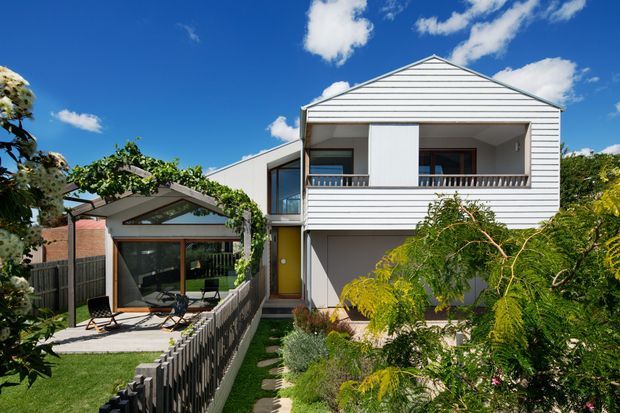 Exterior view of a compact two-storey suburban residence with a white horizontal weatherboard facade and gabled roof. The upper level has a recessed balcony with timber balustrading and dark-framed windows, while the lower level includes a covered entry and garage-like opening. A glazed garden pavilion or sunroom with a pitched roof sits to the left, connected by a narrow landscaped path and enclosed garden. Bright blue sky, established planting, and a small lawn surround the home.