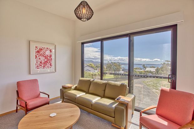A bright living room with a tan leather sofa, two coral upholstered armchairs, a rounded timber coffee table, framed artwork, and a woven pendant light. Large sliding glass doors open to a balcony or terrace with expansive water and landscape views.
