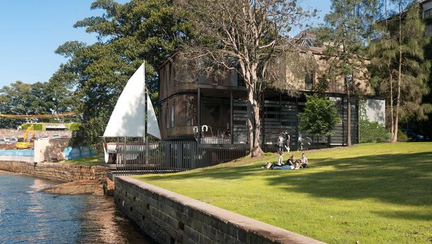 Exterior view of a contemporary multi-level waterfront building beside a calm body of water, with dark timber or metal screening, extensive glazing, a deck and railing, landscaped lawn, stone retaining wall, and a small sailboat in the foreground.
