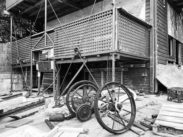 Black-and-white photograph of a raised timber structure under construction or renovation, with a covered upper-level deck enclosed by patterned breeze-block screens. The lower level has exposed brickwork, timber framing, scaffolding, construction materials, and old industrial machinery in the foreground. The image has a documentary, historic construction-site character.