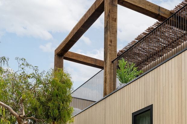 Exterior architectural detail showing a contemporary building facade clad in light vertical weatherboards, with prominent dark timber post-and-beam framing, black metal balustrades, and a slatted overhead screen or pergola. Vegetation and sky surround the structure.