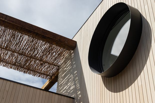 Close-up exterior architectural detail showing a large black oval window projecting from a pale vertical timber-clad facade. A timber pergola or privacy screen with woven reed-like shading casts patterned shadows across the wall, with overcast sky visible above.