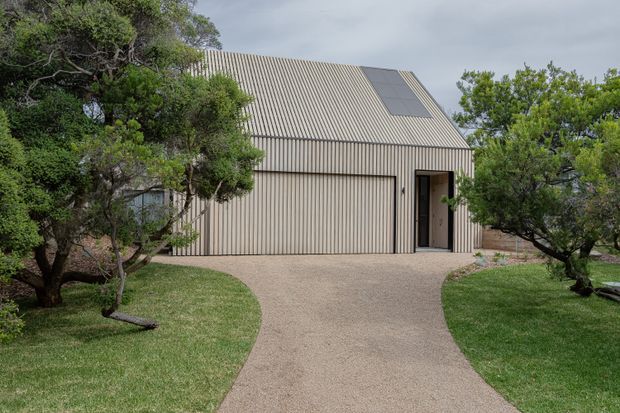 Front exterior view of a contemporary single-storey building with a steep pitched corrugated metal roof and matching vertically ribbed facade. A broad garage door and recessed pedestrian entry face a curved gravel driveway, framed by mature landscaped trees and lawn. Solar panels are visible on the roof.