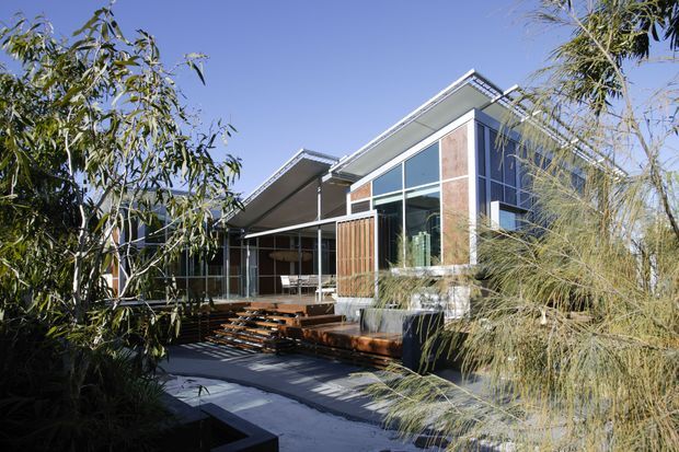 Exterior view of a contemporary single-storey residence set within landscaped planting. The home features angular skillion-style roof forms, extensive floor-to-ceiling glazing, timber-clad wall sections, and a raised timber deck with steps leading to a paved outdoor area.