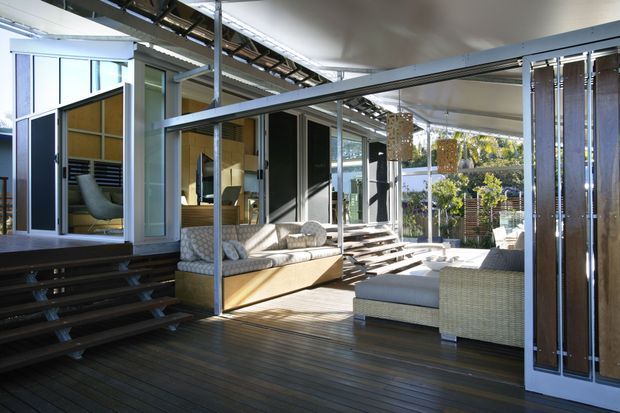 A modern indoor-outdoor living area with extensive glazing, dark timber decking, open sliding or folding glass doors, and a covered terrace. The space includes built-in and freestanding light upholstered seating, a raised platform with steps, exposed structural framing, and vertical timber screening. Strong connection to a landscaped exterior is visible through the open facade.