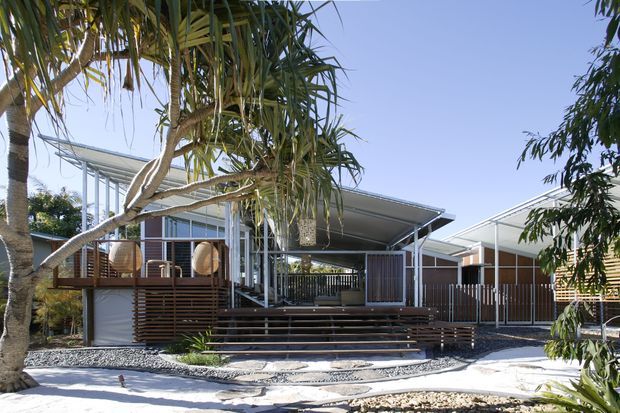 Exterior view of a low-rise contemporary residence with broad skillion-style roof planes, extensive glazing, timber screening, elevated timber decking and a stepped entry terrace. The home is set within a landscaped garden with tropical planting and exposed gravel landscaping.