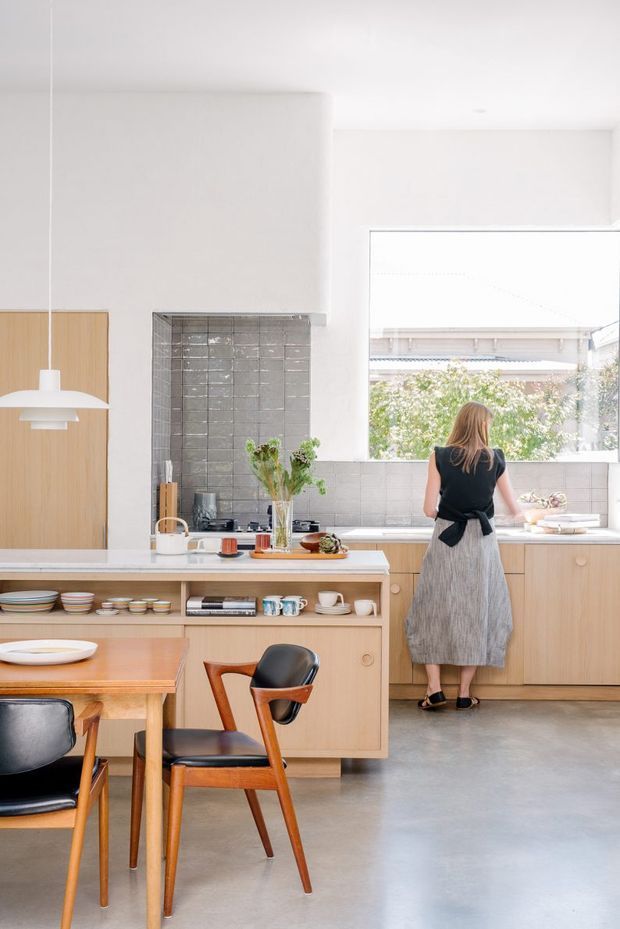 Bright, open-plan kitchen and dining area with light timber cabinetry, a central island with open shelving, grey tiled splashback, polished concrete flooring, and a large rear window. A warm timber dining table and mid-century style chairs occupy the foreground, while a person stands at the kitchen bench. The space has a clean, airy, understated contemporary character.