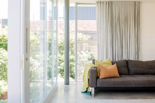Bright contemporary living room with full-height floor-to-ceiling glazing, slim white frames, exposed cylindrical structural columns, pale polished concrete flooring, and a dark charcoal sofa with yellow and orange cushions. Vertical sheer curtains soften the glazing, while a brick building and greenery are visible outside.