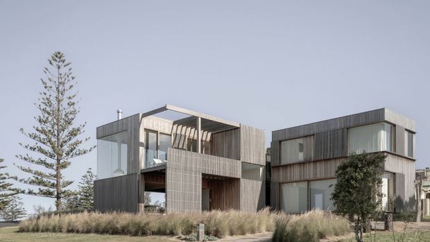 Exterior view of a contemporary multi-volume residential building composed of two connected box-like forms. The facade features extensive floor-to-ceiling glazing, vertical timber-like cladding, projecting upper levels, recessed balconies, and flat roofs. Coastal-style grasses and a tall conifer frame the foreground, with a pale sky and hints of neighbouring buildings in the background.