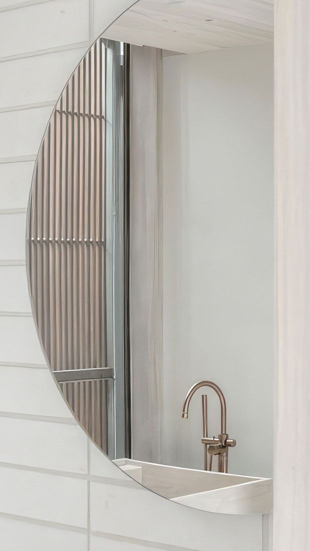A close-up view of a refined bathroom vanity area, dominated by a large round mirror reflecting vertical decorative screening and a pale timber-lined ceiling or wall. A white basin with a slim metallic gooseneck tap sits below the mirror, with light neutral surfaces and subtle curved detailing throughout.