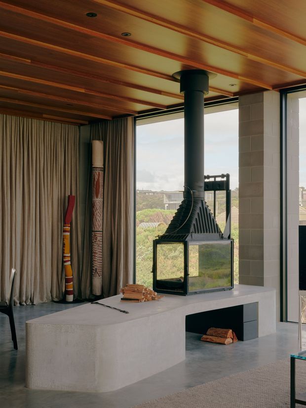 Interior view of a contemporary living area centred on a sculptural black wood-burning fireplace with a glass firebox, set on a large pale plaster or concrete hearth. The room features a warm timber-lined ceiling with recessed downlights, full-height pleated curtains, polished grey concrete flooring, light blockwork walls, and expansive glazing overlooking the landscape. Decorative objects lean against the curtained wall, while a small portion of seating and a rug are visible at the edges.