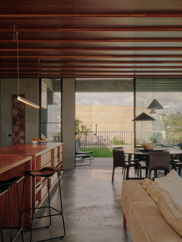 A contemporary open-plan interior combining kitchen, dining and living areas. The room features a long timber kitchen island with integrated cabinetry and dark bar stools, a polished concrete floor, exposed timber ceiling beams, dark tiled wall surfaces, and expansive floor-to-ceiling glazing opening to a balcony or terrace. A dining table with pendant lights and a neutral sofa occupy the adjoining space, creating a warm, refined indoor-outdoor connection.