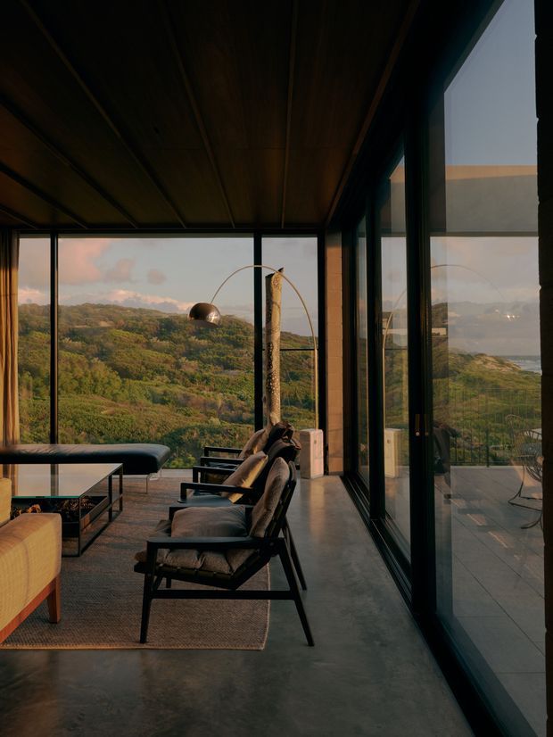 A refined modern living room with expansive floor-to-ceiling glazing overlooking a lush, hilly landscape and distant coastline. The room features a dark timber-lined ceiling, polished concrete floor, black-framed glass walls, a low curved sofa, sculptural lounge chairs, a glass coffee table, and a large arched floor lamp. Warm evening light creates a calm, luxurious atmosphere.