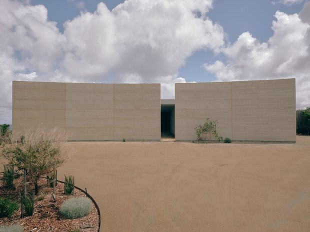 A minimalist single-storey building viewed from the front, composed of two long, rectangular pale beige masonry or rendered volumes separated by a narrow dark recessed entrance. The building sits within a broad sandy forecourt with sparse drought-tolerant landscaping, including succulents and low shrubs. The flat roofline, monolithic walls, restrained openings, and warm desert-like setting create a stark contemporary architectural composition beneath a partly cloudy sky.