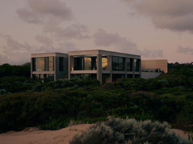 A contemporary two-storey residence set amid dense coastal scrub and sandy dunes. The building has a low, rectilinear form with extensive dark-framed glazing, exposed concrete or grey rendered structural elements, projecting upper-level volumes, and a flat roof. The image is captured at dusk under a cloudy sky, creating a subdued, moody atmosphere.