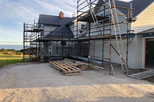 Exterior view of a multi-part weatherboard-clad building under construction or renovation, with dark pitched metal roofs, scaffolding, exposed decking materials, and an attached garage or service opening. The property overlooks open coastal or rural landscape.