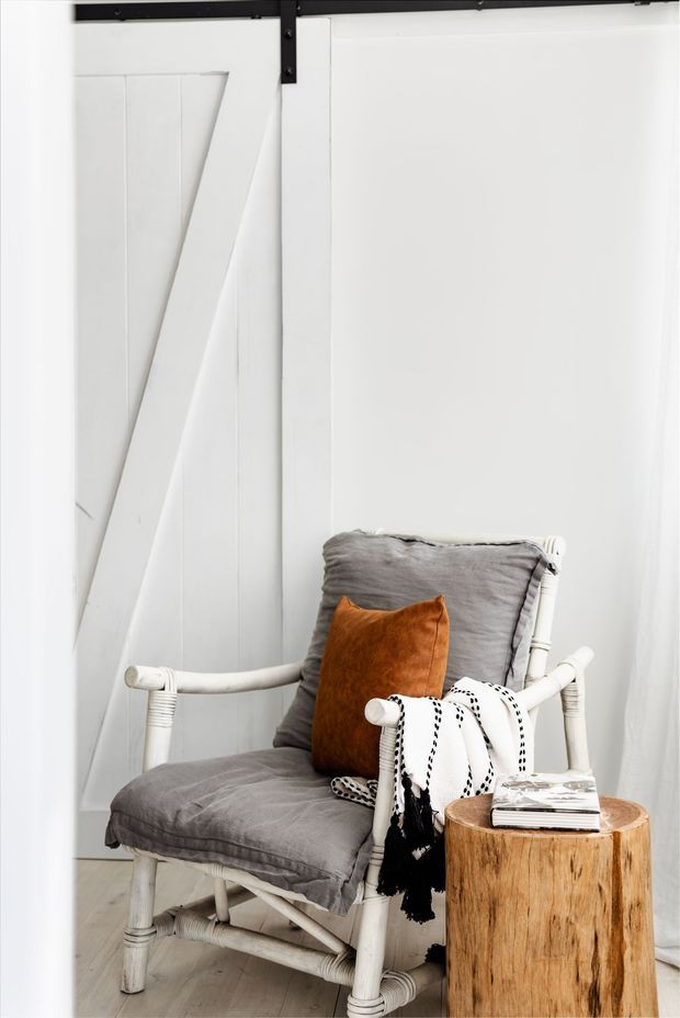 A bright, white-toned interior corner featuring a white painted sliding barn door with black metal hardware, a white woven or rattan-style armchair with grey cushions, and a small natural timber stump side table. An orange-brown cushion and patterned throw add warmth and contrast. The styling is simple, rustic, and relaxed.