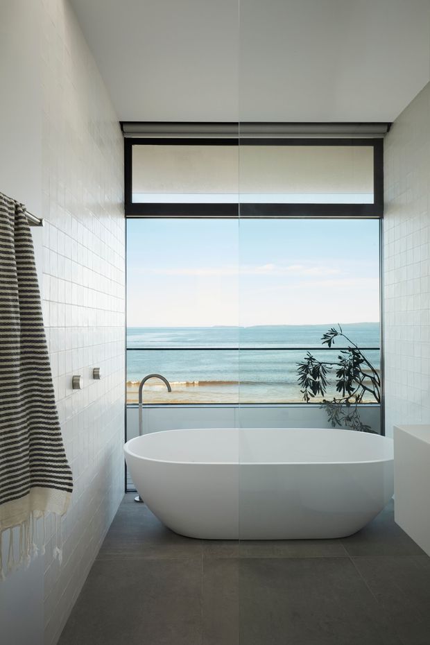 A bright, narrow contemporary bathroom with a freestanding white bathtub positioned in front of full-height glazing overlooking the ocean. White tiled walls, dark grey floor tiles, black-framed upper and lower glazing, a wall-mounted towel rail with a striped towel, and a slim bath filler are visible. The space has a clean, calm coastal atmosphere and strong indoor-outdoor connection.