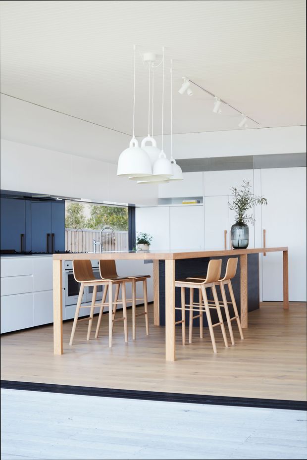 A bright, open-plan contemporary kitchen viewed from an adjoining area. The space features white full-height cabinetry, a dark navy kitchen wall and island face, pale timber flooring, and a long timber dining-height island or table with integrated seating. Several light timber bar stools surround the island. White pendant lights hang above the island, with additional white track lighting on the ceiling. Large glazing or an open connection at the rear brings in outdoor light and views.