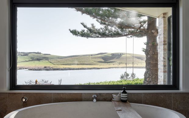 A freestanding bathtub is positioned directly beneath a large black-framed picture window, overlooking a calm body of water and rolling green hills. The bathroom features dark stone-look wall tiling around the tub, a pale tub, and a timber-toned ledge with minimal bath accessories. A mature tree and hanging swing are visible outside.