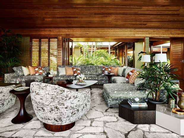 A richly layered indoor living room with a low timber-lined ceiling and walls, extensive louvre and glazed openings, patterned sectional seating, sculptural lounge chairs, dark wood side tables, indoor tropical plants, and a pale irregular stone floor. The room has a relaxed, resort-like atmosphere with strong mid-century and tropical influences.