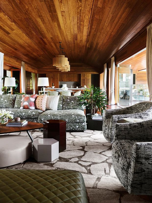 A richly styled living room with extensive warm timber panelling across the ceiling and walls, floor-to-ceiling glazing with curtains, a patterned sectional sofa, textured armchairs, layered coffee tables, indoor plants, and a sculptural pendant light. The room opens visually toward a dining or outdoor area through large glass doors. The palette is warm, earthy, and organic with bold botanical upholstery and a geometric rug.
